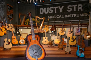 An array of fretted instruments, plus a harp and a hammered dulcimer, on a low wooden stage with a resonator guitar in the forefront and Dusty Strings sign on the wall