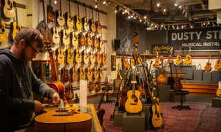Tall room with warm lighting and guitars covering every wall and surface. Someone plays guitar on a low stage and someone else restrings a guitar on a workbench.