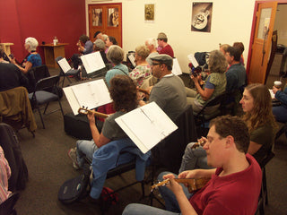 Group of people playing instruments in a classroom setting with music stands and sheet music.